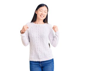 Genç, güzel, Çinli bir kadın gündelik süveter giyiyor. Çok mutlu ve heyecanlı. Kolları havada, gülümseyerek ve başarı için çığlık atarak kazanan bir jest yapıyor. kutlama konsepti. 