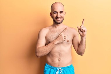 Mayolu, kel ve yakışıklı bir adam. Gülümsüyor ve kameraya bakıyor. İki eliyle ve parmaklarıyla yan tarafa bakıyor.. 