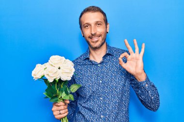 Çiçek tutan yakışıklı genç adam parmaklarıyla imza atıyor, gülümseyerek mükemmel bir sembol oluşturuyor. 