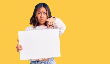 Genç, güzel, melez bir kadın elinde boş bir pankartla kamerayı ve seni işaret eden kendine güvenen, ciddi görünen bir jest. 