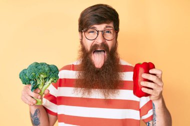 Uzun sakallı, elinde brokoli ve kırmızı biber olan yakışıklı, genç, kızıl saçlı, komik bir ifadeyle dilini dışarı çıkaran adam.. 