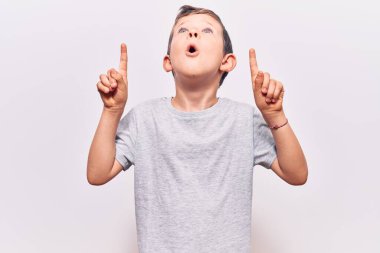 Gündelik kıyafetler giyen sevimli sarışın çocuk şaşırmış ve şaşırmış bir şekilde yukarı bakıyor ve parmaklarıyla işaret ediyor ve kollarını kaldırıyor.. 