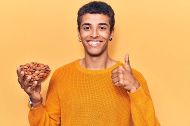 Genç Afrikalı Amerikalı elinde bademlerle gülümseyen mutlu ve pozitif bir adam, başparmağını kaldırarak mükemmel ve onay işareti yapıyor. 