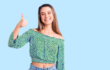 Gündelik tişört giyen genç ve güzel bir kız mutlu ve pozitif gülümsüyor, başparmağıyla mükemmel ve onay işareti yapıyor. 