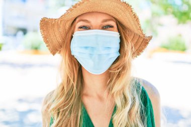 Yaz şapkası ve tıbbi maske takan, tatilde olan genç, güzel sarışın kadın. Şehrin caddesinde yüzünde gülümsemeyle dikilirken.