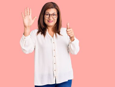 Orta yaşlı Latin kadın gündelik kıyafetler ve gözlük takıyor. Altıncı parmaklarını gösterip işaret ederken kendinden emin ve mutlu gülümsüyor.. 