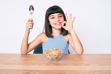 Bangır bangır küçük bir kız masada oturmuş mısır gevreği yiyor parmaklarıyla işaret veriyor, gülümsüyor ve mükemmel bir sembol oluşturuyor. 