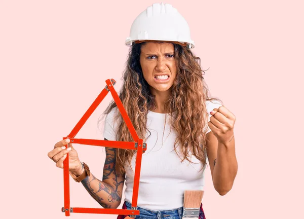 Young hispanic woman with tattoo wearing architect hardhat holding build project annoyed and frustrated shouting with anger, yelling crazy with anger and hand raised