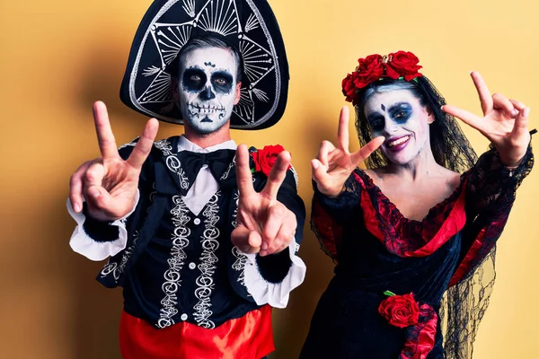黄色の笑みを浮かべて舌の上に死んだ衣装のメキシコの日を着て若いカップルの両手の勝利のサインを行うの指を示しています — ストック写真