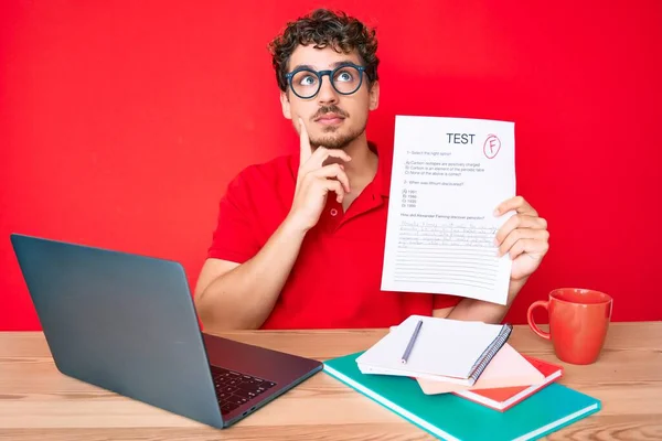 Young caucasian man with curly hair sitting on the table showing failed exam serious face thinking about question with hand on chin, thoughtful about confusing idea