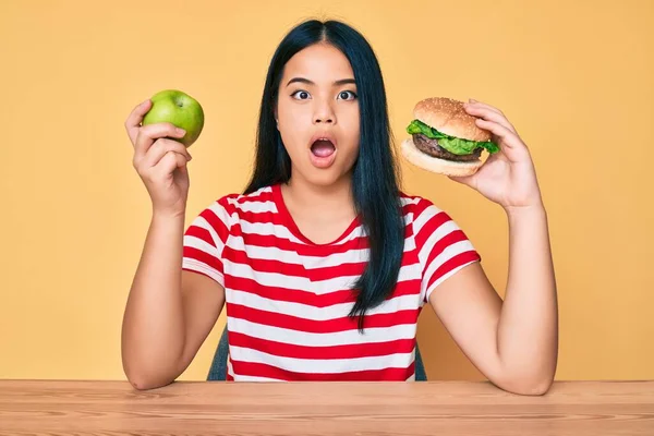 年轻的亚洲女孩比较汉堡包和健康的苹果 惊讶的表情 恐惧和兴奋的脸 — 图库照片
