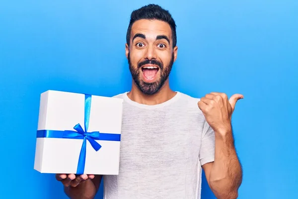 Jovem Hispânico Homem Segurando Presente Apontando Polegar Até Lado Sorrindo — Fotografia de Stock