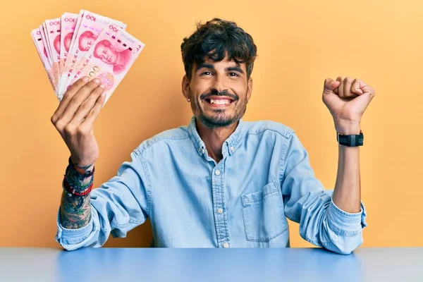 Jovem Hispânico Segurando Notas Chinesas Yuan Sentado Mesa Gritando Orgulhoso — Fotografia de Stock