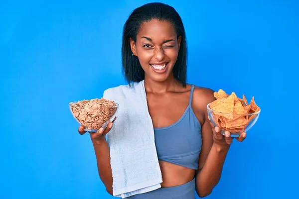 身穿运动服 拿着玉米片和麦片的年轻的非洲裔美国女人 带着性感的表情 欢快而快乐的脸看着摄像机 眨了眨眼 — 图库照片