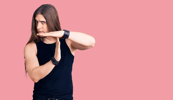 Young adult man with long hair wearing goth style with black clothes doing time out gesture with hands, frustrated and serious face