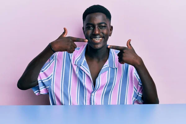 若いアフリカ系アメリカ人の男性は テーブルの上に座っているカジュアルな服を着て明るい表情と指の歯と口で指して笑顔 歯の健康の概念 — ストック写真