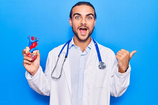 Jovem Homem Bonito Vestindo Estetoscópio Médico Segurando Coração Apontando Polegar — Fotografia de Stock