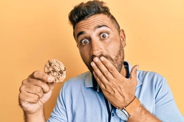 Sakallı yakışıklı adam elinde kurabiyeyle ağzını eliyle kapatıyor, şokta ve hata yapmaktan korkuyor. şaşırmış ifade 