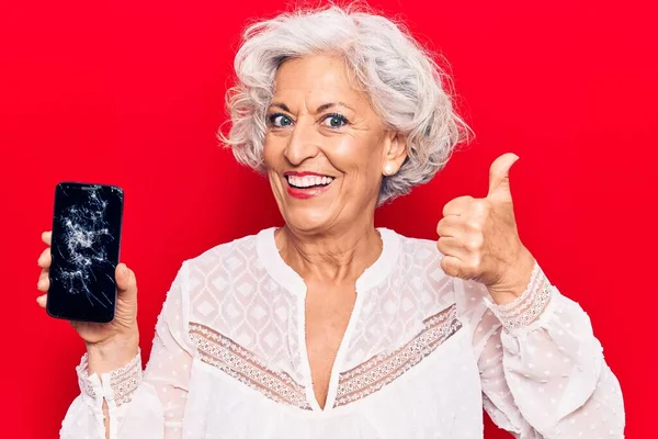 Senior grey-haired woman holding broken smartphone showing cracked screen smiling happy and positive, thumb up doing excellent and approval sign