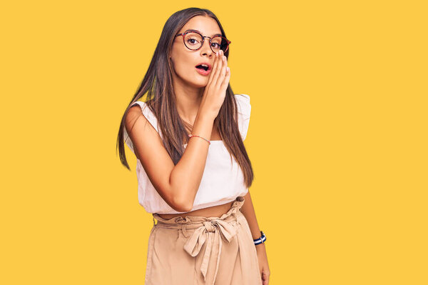 Young hispanic woman wearing casual clothes and glasses hand on mouth telling secret rumor, whispering malicious talk conversation 
