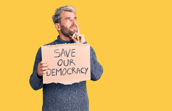 Young blond man holding save our democracy cardboard banner serious face thinking about question with hand on chin, thoughtful about confusing idea 