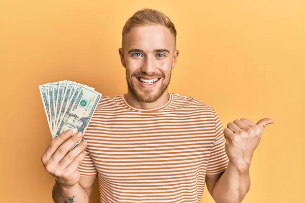 Joven Hombre Caucásico Sosteniendo Dólares Señalando Pulgar Hacia Lado Sonriendo —  Fotos de Stock