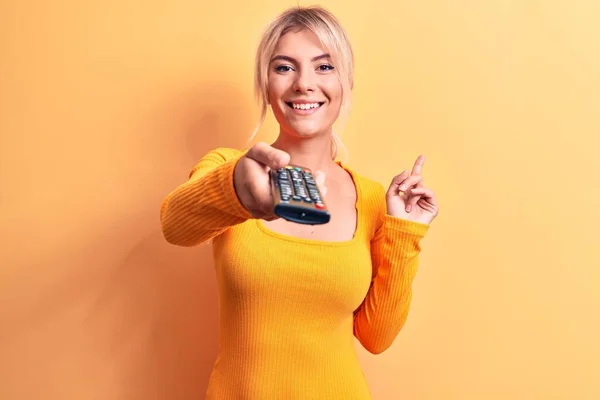 金发女人换电视频道用电视遥控装置在黄色背景上开心地微笑着 手指头指向旁边 — 图库照片