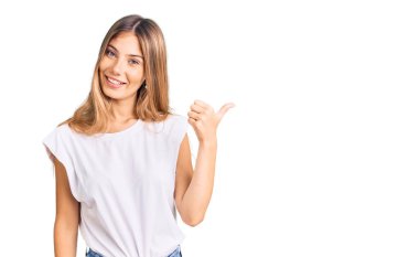 Sarışın, beyaz tişörtlü, neşeli yüzlü, gülümseyen ve baş parmağıyla yüzünü gösteren güzel beyaz bir kadın.. 