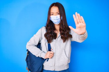 Genç, güzel Çinli kız, öğrenci çantası tutuyor, tıbbi maske takıyor ve elinde dur işareti var, ciddi ve kendinden emin bir ifadeyle, savunma hareketi yapıyor. 