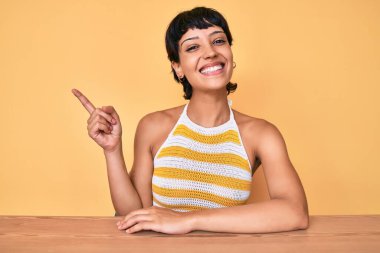 Masada oturan güzel esmer kadın sarı arka planda gülümsüyor mutlu bir şekilde elini ve parmağını yana uzatıyordu. 