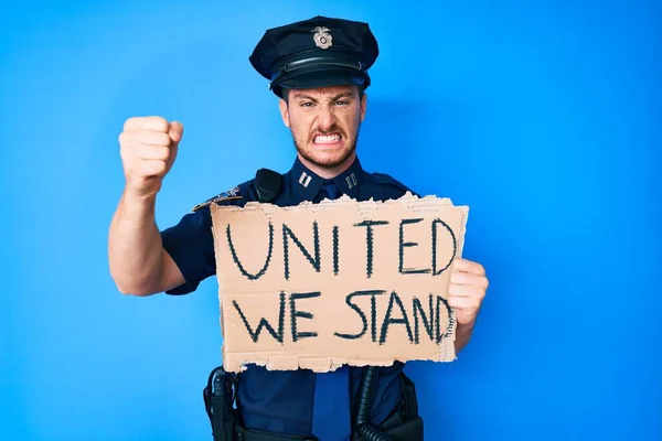 Young caucasian man wearing police uniform holding united we stand banner annoyed and frustrated shouting with anger, yelling crazy with anger and hand raised