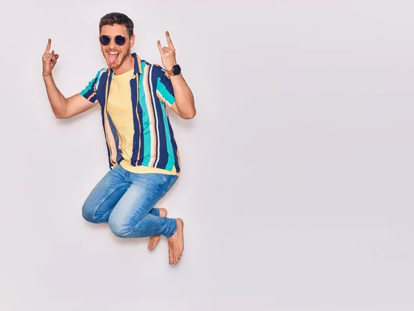 カジュアルな服とサングラスを身に着けている若いハンサムなヒスパニック系の男は幸せ笑う 白い背景の上に指で角記号を行う顔に笑顔でジャンプ — ストック写真
