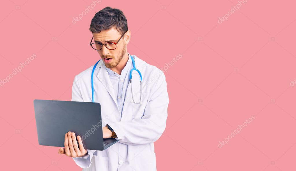 Handsome young man with bear wearing doctor uniform working using computer laptop scared and amazed with open mouth for surprise, disbelief face 