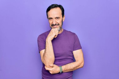 Orta yaşlı, mor tişört giyen, izole edilmiş arka planda duran yakışıklı adam kameraya kollarını kavuşturup çenesine dokunarak gülümsüyor. Olumlu düşünüyorum..