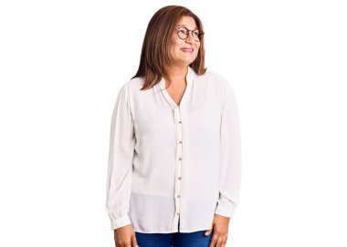 Orta yaşlı, sıradan kıyafetler ve gözlük takan, yüzünde gülümseme, doğal bir ifade olan Latin bir kadın. Kendinden emin gülen. 