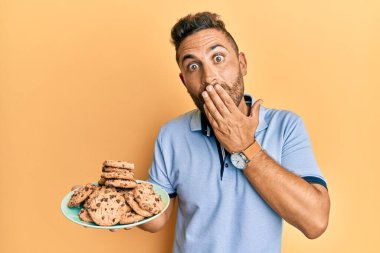 Sakallı yakışıklı adam elinde kurabiyelerle ağzını eliyle kapatıyor, şokta ve hata yapmaktan korkuyor. şaşırmış ifade 