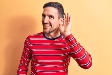 Sıradan çizgili tişört giyen genç yakışıklı adam izole edilmiş sarı arka planda dikiliyor. Kulaktan kulağa gülümsüyor ve söylenti ya da dedikoduları dinliyor. Sağırlık kavramı.