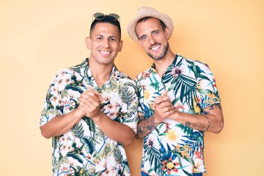 Yazlık şapkalı, Hawaii tişörtlü, elleri çapraz parmaklı rahat ve neşeli gülümseyen iki genç eşcinsel. Başarı ve iyimserlik 