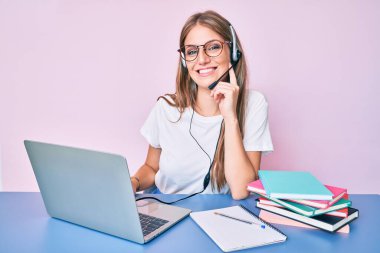 Çağrı merkezindeki operatör kulaklığı takan genç sarışın kız kameraya kollarını kavuşturmuş ve çenesine dokunmuş bir şekilde gülümsüyor. Olumlu düşünmek. 