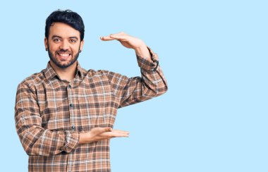 Latin asıllı genç adam, sıradan kıyafetler giyiyor. Elleriyle işaret ediyor. Ölçü sembolü, büyük boy işareti. Kameraya bakıp gülümsüyor. ölçüm kavramı. 