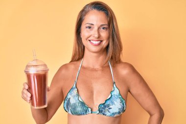 Orta yaşlı İspanyol kadın elinde bir bardak smoothie ile pozitif ve mutlu görünüyor ve kendine güvenen bir gülümsemeyle dişlerini gösteriyor. 