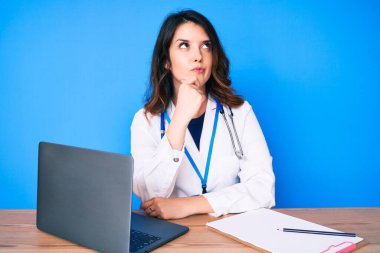 Genç, güzel esmer kadın doktor üniforması giyiyor klinikte çalışıyor şüphe üzerine yoğunlaşmış düşünüyor çene üzerinde parmak ve merak içinde yukarı bakıyor 