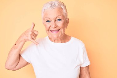 Son sınıf güzeli, mavi gözlü, gri saçlı, sarı arka planda klasik beyaz tişört giyen, telefonla el ve parmak hareketleriyle gülümseyen, telefonda konuşan bir kadın. iletişim kavramları. 