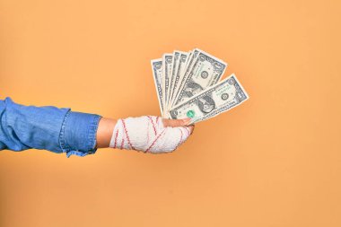 Kafkasyalı genç bir kadının yaralı eli, sargıyla kaplı, izole edilmiş sarı arka planda bir deste banknot tutuyor.