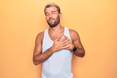 Sarışın, genç bir adam izole edilmiş sarı arka planda kolsuz tişört giyiyordu. Elleri göğsünde gülümsüyordu. Yüzü minnettarlıkla kapalıydı. Sağlık kavramı.
