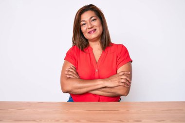 Orta yaşlı esmer, sıradan kıyafetler giyen, masanın üzerinde oturan mutlu yüzlü, kollarını kavuşturup kameraya bakan bir kadın. pozitif kişi. 