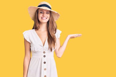 Genç, sarışın, yaz elbisesi ve şapkası giymiş neşeli bir şekilde gülümseyen, avuç içiyle kameraya bakan güzel bir kadın.. 