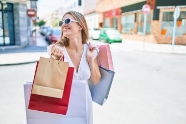 Genç sarışın kadın gülümsüyor. Şehrin caddesinde alışveriş torbalarını tutarken mutlu.