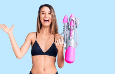 Bikini giyen güzel bir kız elinde su tabancasıyla zaferi kutluyor mutlu bir gülümsemeyle ve kazanan bir ifadeyle ellerini kaldırarak. 