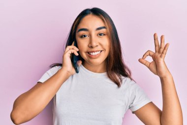 Genç Latin bir kadın akıllı telefonda konuşuyor parmaklarıyla imza atıyor, gülümsüyor ve mükemmel bir sembol oluşturuyor. 